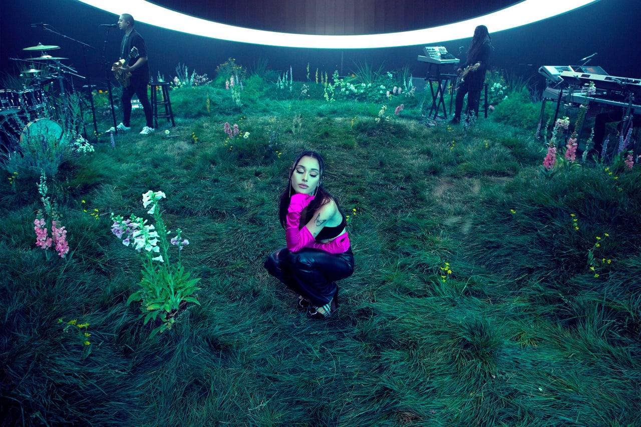 Ariana performs in an indoor meadow beneath a glowing oval light, with instruments behind her. Flowers, foliage and keyboards extend to both edges. Source: WallpaperCat (uploader sonny.gutmann). Original artist not identified.