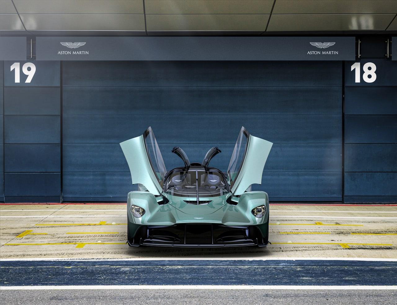 A Valkyrie faces forward with raised doors between numbered pit garages. Painted lane markings and the garage fronts give the vehicle a restrained architectural setting. Source: 4kwallpapers.com. Original artist not identified by the source page.