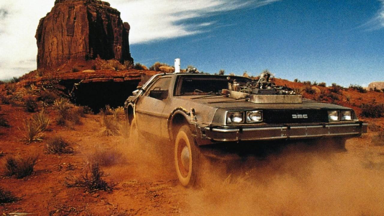 The DeLorean races through red desert dust beside a tall rock formation. Scrub, mesa walls and open terrain extend beyond the time machine.