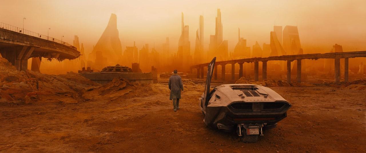 A lone figure and spinner enter a dust-orange city of broken roads and distant towers. Elevated structures and rubble continue across both edges of the wide film still.