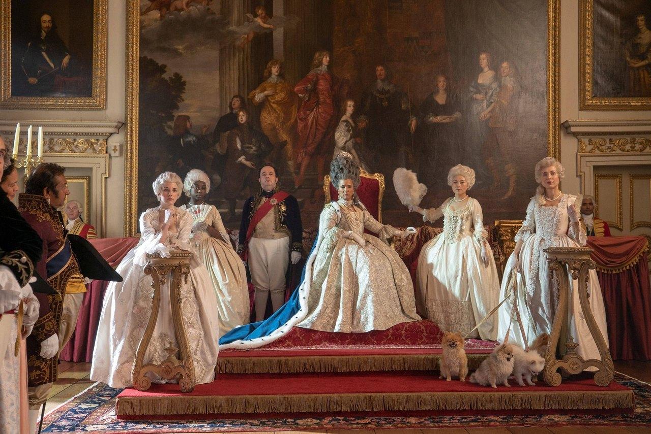 A line of elegantly dressed women and attendants stands in an ornate room filled with portraits, gilding and red upholstery.
