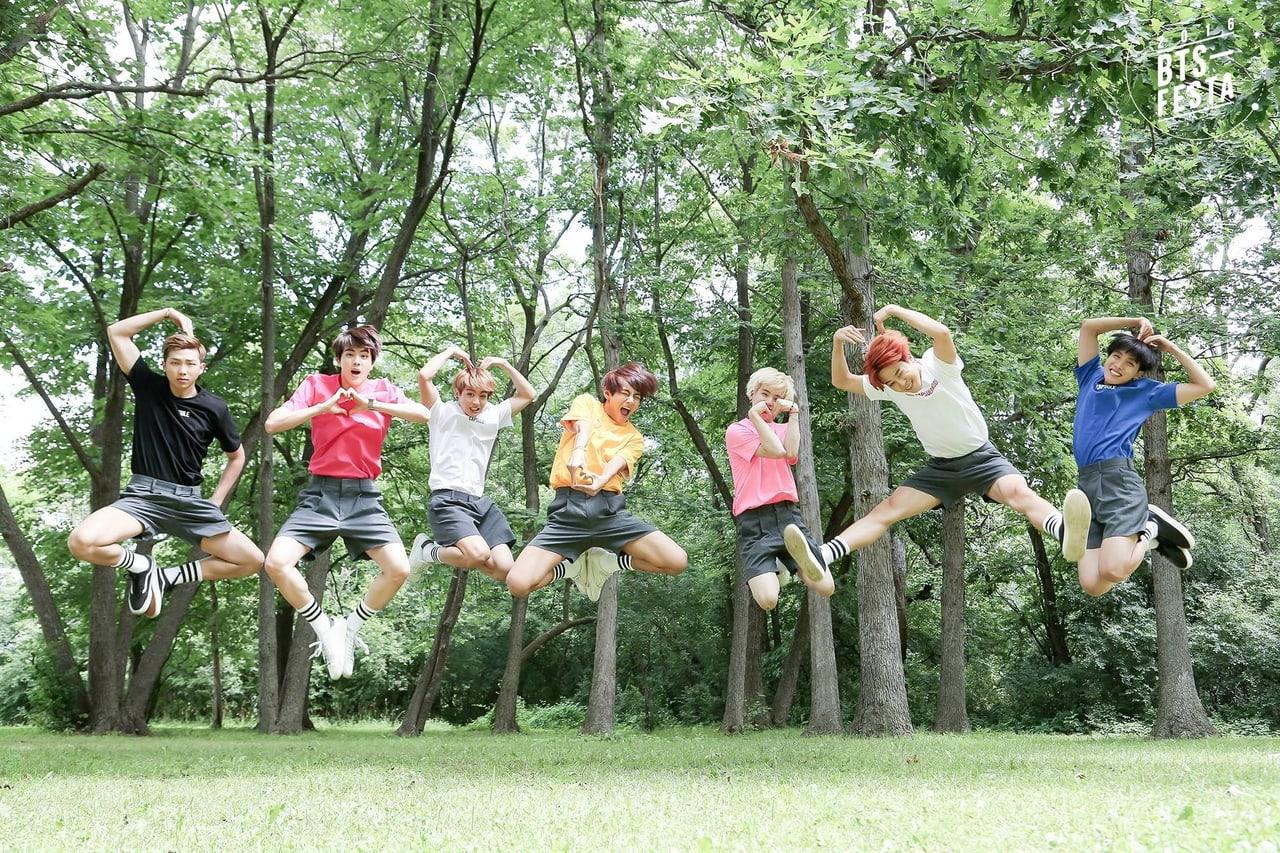 Seven members jump in a line among green trees above a grassy clearing.