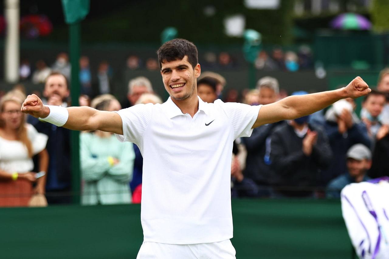 Alcaraz celebrates with open arms in traditional white tennis clothing. Deep green court surroundings and spectators create a quieter photographic backdrop. Source: struggle on Wallpapers.com. Original artist not identified by the page.