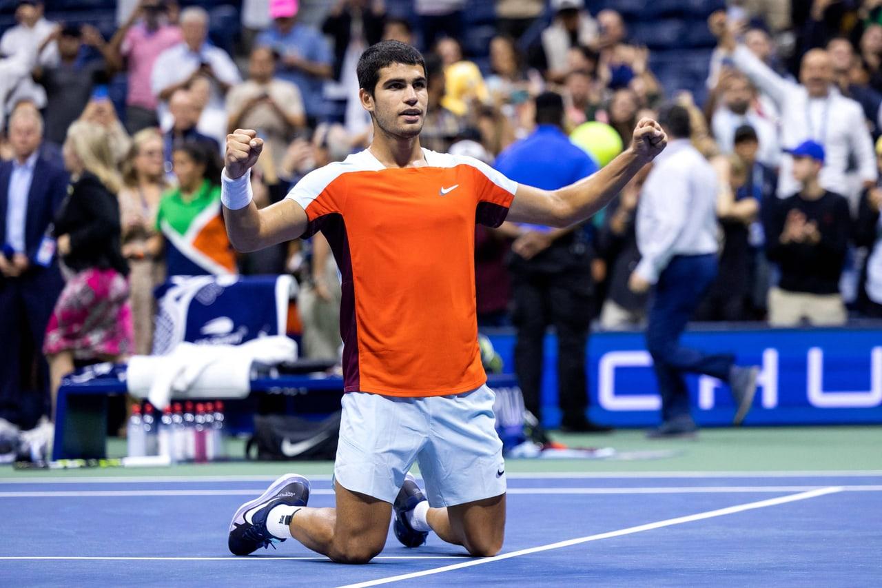Alcaraz kneels on a blue hard court in an orange-and-white shirt, arms raised toward the stands. The crowd and court markings fill the wider scene. Source: e2fq7fzj on Wallpapers.com. Original artist not identified by the page.