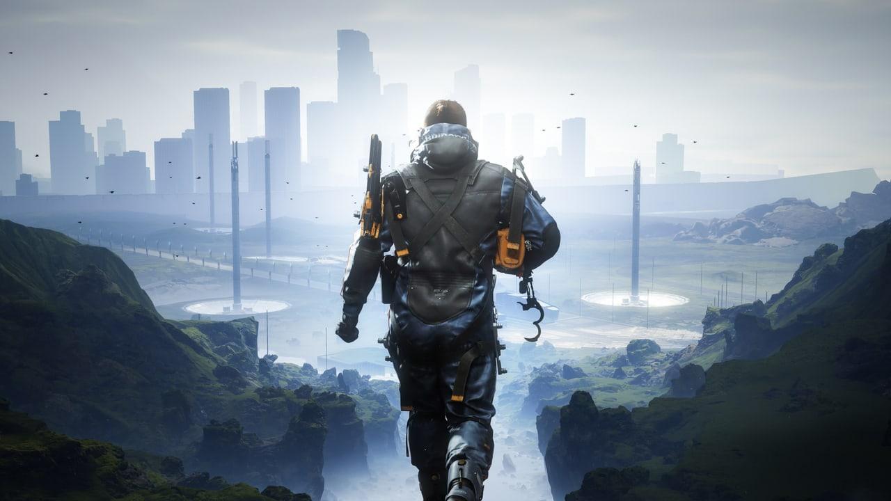 Sam walks through a broad rocky valley toward pale futuristic buildings. Cliffs, green ground cover and distant structures provide depth beyond the central figure.