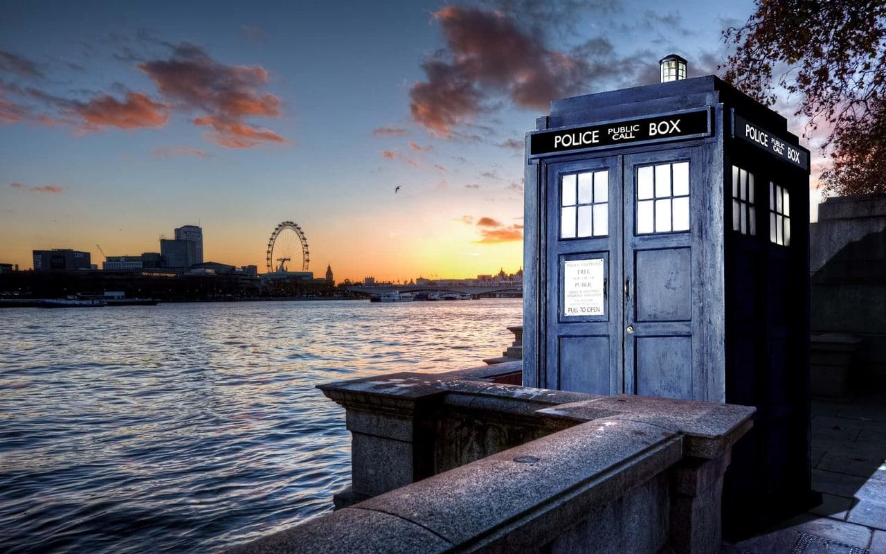 The blue police box stands on a riverside terrace at sunset. London buildings and water fill the left, while the box, stone railing and tree canopy hold the right.