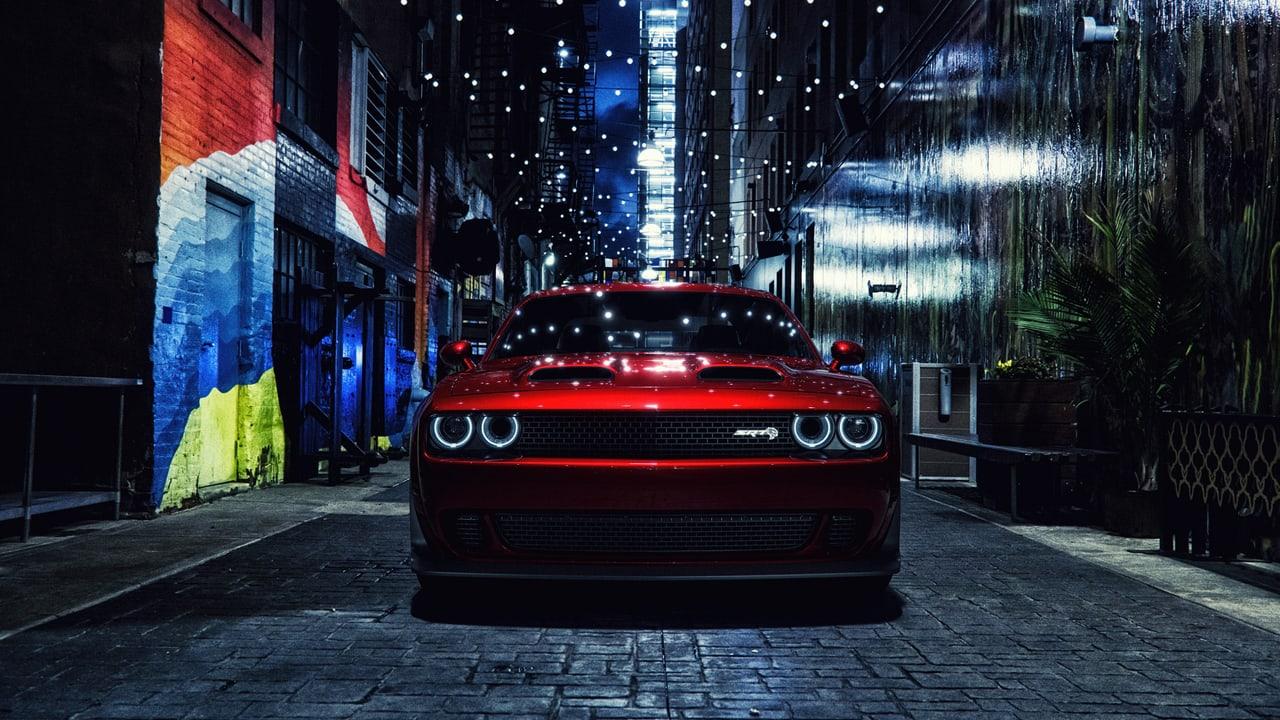 A red Challenger occupies a narrow night alley under strings of lights. Painted brick, glossy walls, foliage and cobblestones frame both sides.