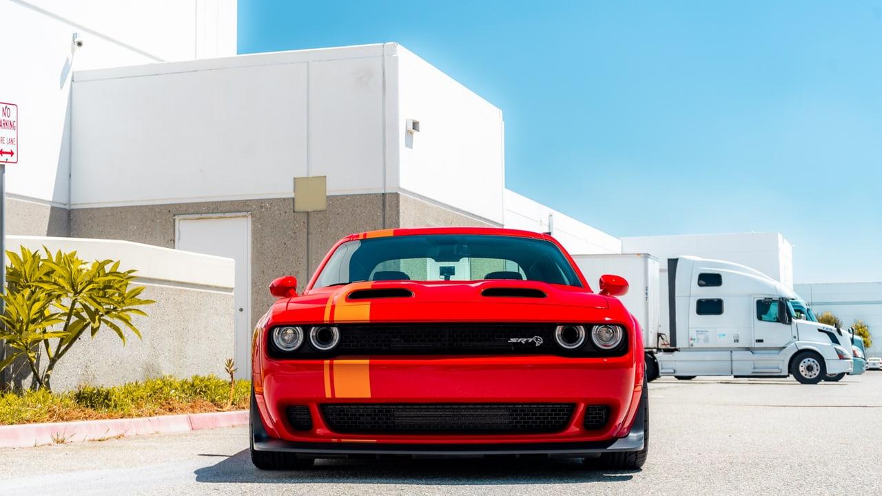 A red Challenger faces forward outside a white industrial building. A small tree and facade fill the left, while parked trucks establish the right edge.