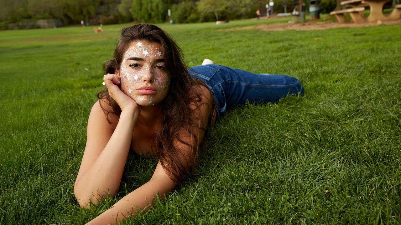 Dua Lipa reclines on a green lawn with small flowers around her face. The garden, grass texture and distant benches give the portrait an outdoor setting. Source: 4kwallpapers.com. Original artist not identified by the source page.