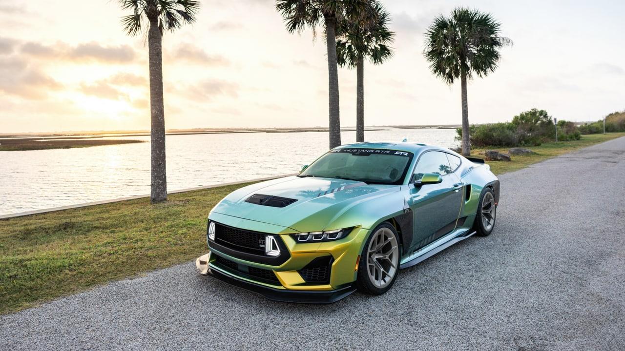 A reflective Mustang stands on a palm-lined waterfront road. Grass and water fill the left while the road and shoreline vegetation continue to the right.