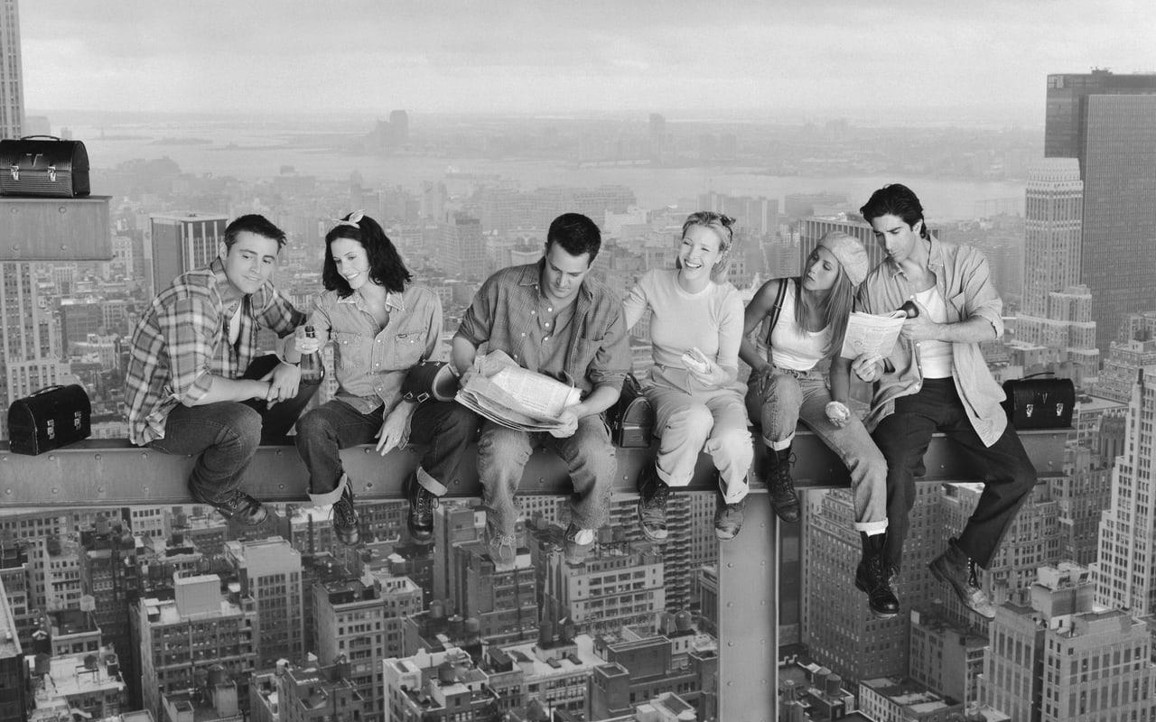 The Friends cast sits on a steel beam above a monochrome Manhattan skyline. Buildings and lunch bags frame both ends of the elevated group portrait.