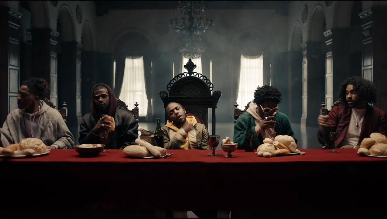 Kendrick Lamar sits at the center of a long red-covered table in a vaulted room. Guests, bread and dark columns extend beyond the central throne into both margins.
