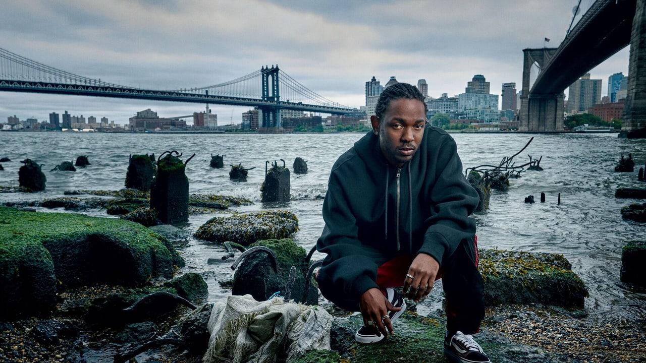 Kendrick Lamar sits on a rocky shoreline between two bridges. Moss-covered posts and suspension cables fill the left while bridge supports and city buildings frame the right.
