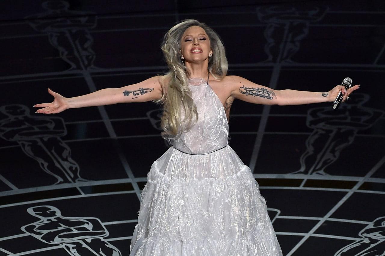Lady Gaga opens her arms on a dark stage patterned with line-drawn statuettes. Her hands and the repeating stage motifs extend into both side strips.
