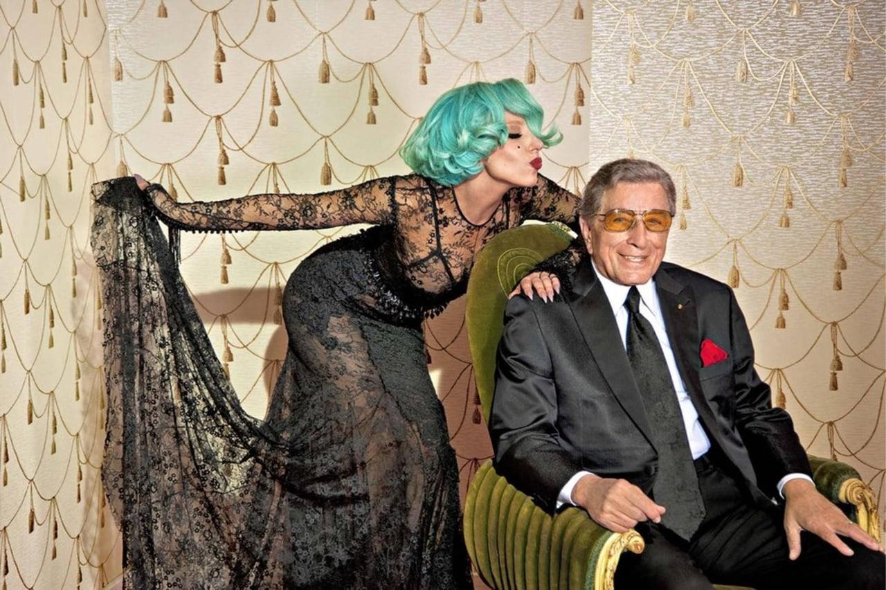 Lady Gaga leans toward Tony Bennett in an ornate room. Black lace on the left and a gold patterned wall and armchair on the right frame the pair.