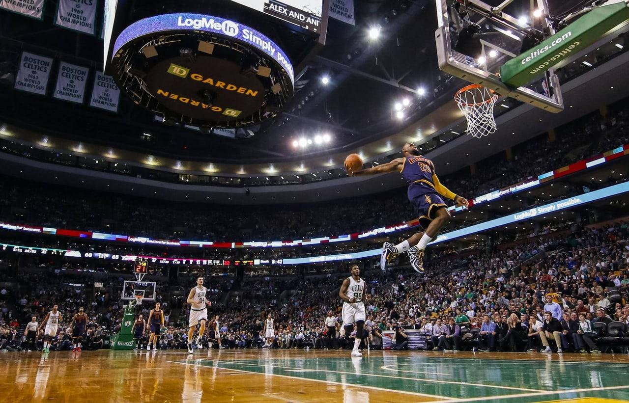 LeBron rises toward the basket in a Lakers uniform inside a packed arena. The hardwood, crowd and roof structure fill a wide view of the dunk. Source: tatart1323 on Wallpapers.com. Original artist not identified by the page.