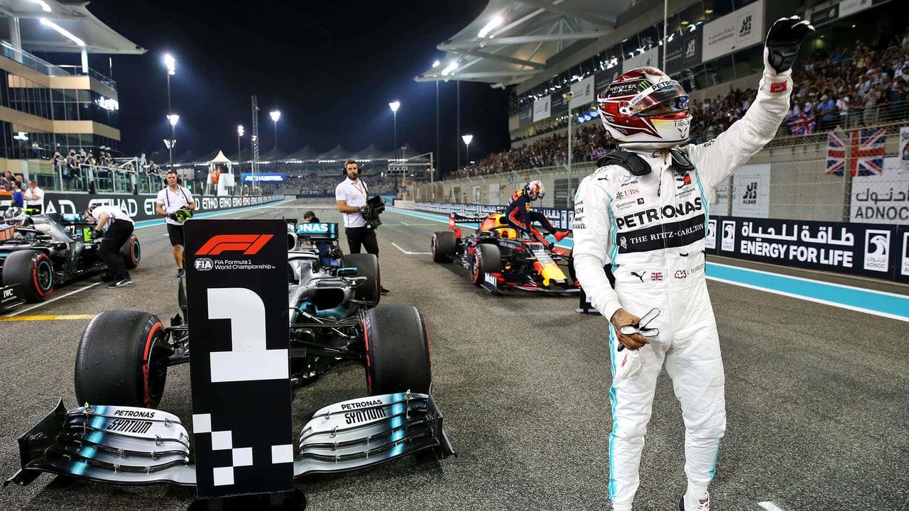 Lewis Hamilton raises a fist on the right of a floodlit Formula One grid with his Mercedes, a number-one board and pit activity across the left.