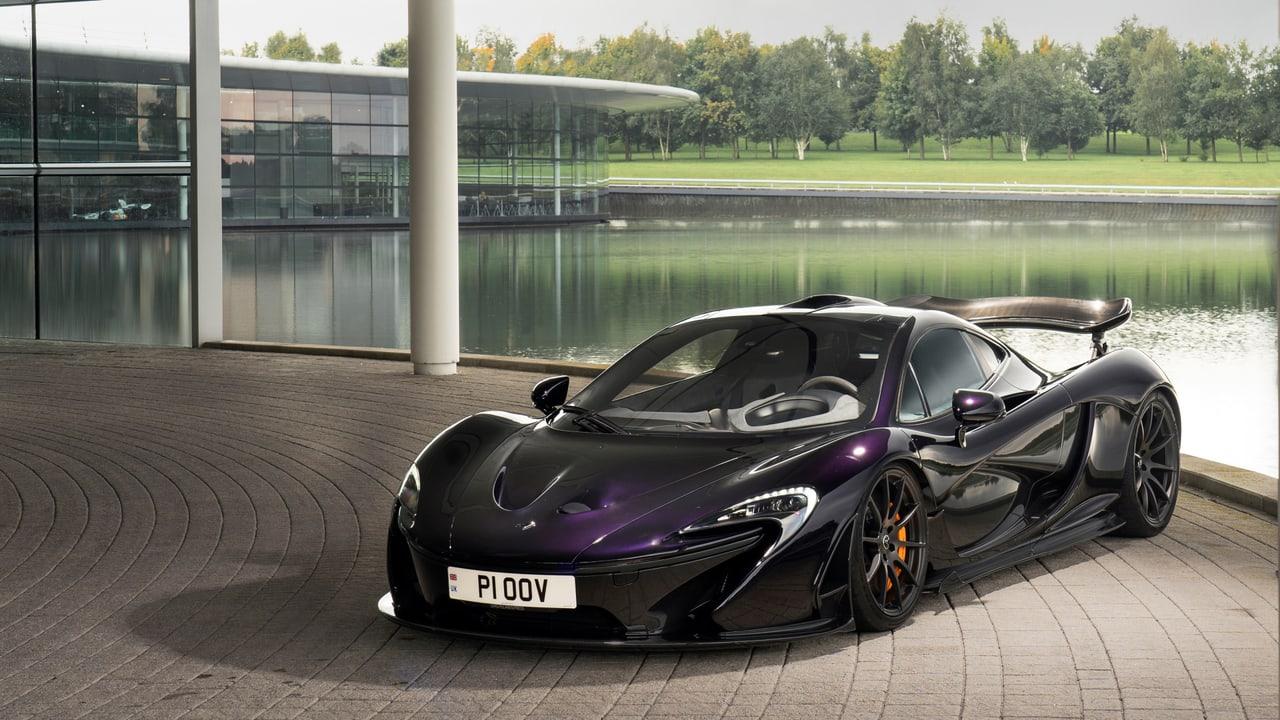 A dark purple McLaren P1 sits on circular paving beside glass architecture and a bright lake.