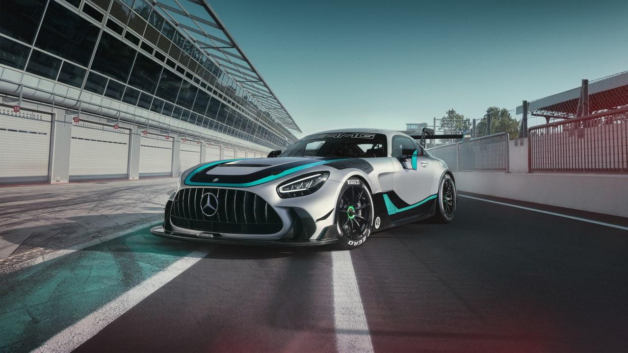 An AMG GT race car rests beside long glass pit buildings and red fencing. Garage doors and converging track markings carry the scene around the conversation.