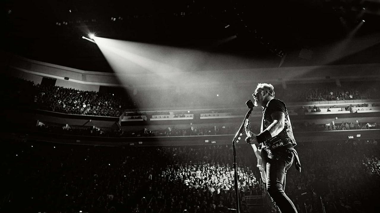 James Hetfield performs in a monochrome arena photograph. The audience and layered seating remain visible at the edges beneath a diagonal white spotlight.