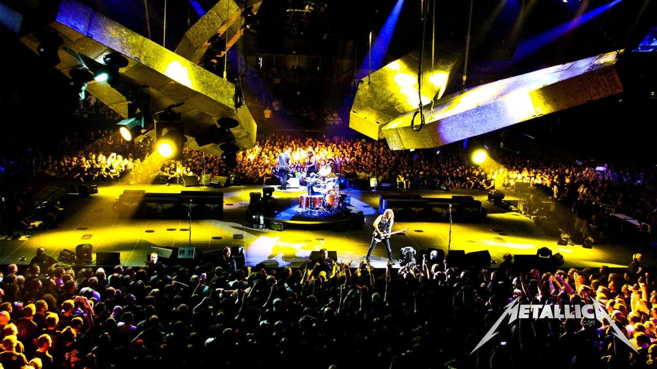 A Metallica arena concert spreads beneath large suspended screens and yellow spotlights. Fans fill the lower frame while rigging and stage displays extend into both sides.