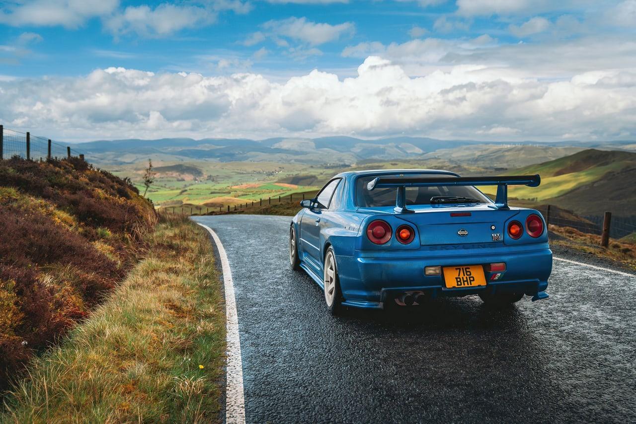 A blue R34 stands on a winding road among rolling hills beneath a partly cloudy sky.