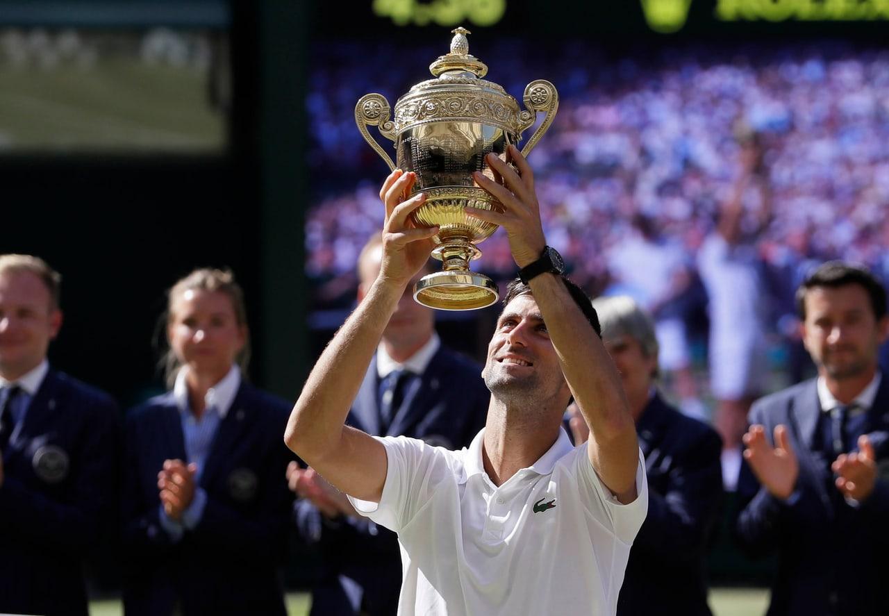 Djokovic raises the Wimbledon trophy before applauding officials and spectators. Figures at both edges retain the ceremony around the central silver-gilt cup.