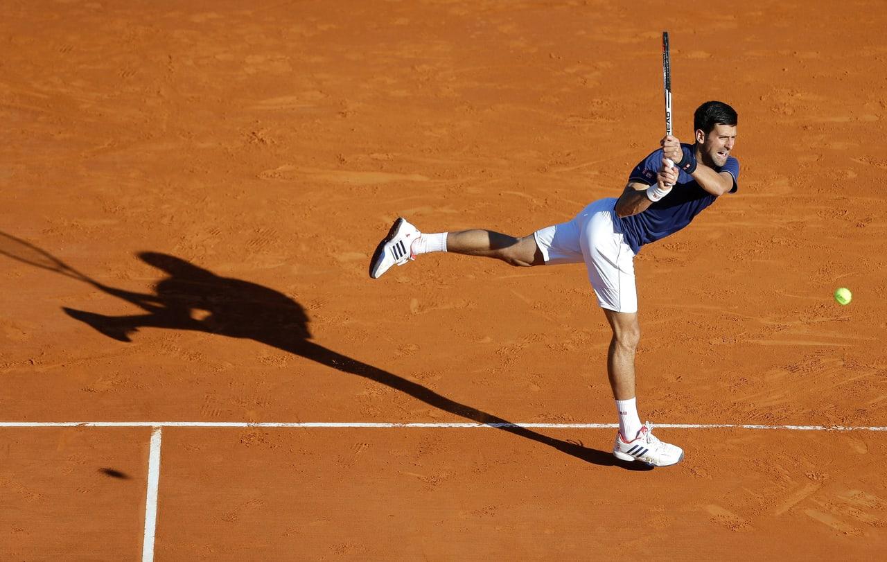 Novak Djokovic follows through on a clay court under strong sunshine. His long shadow reaches left, while a tennis ball and marked court surface remain on the right.