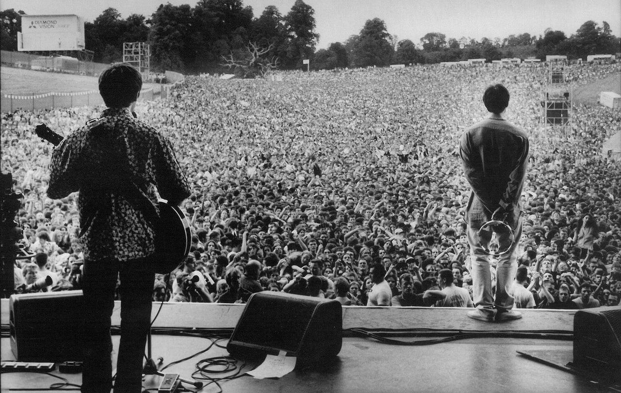 A monochrome Oasis concert photograph looks from the stage toward a vast audience. A guitarist, monitors, speaker towers and thousands of fans stretch across the scene.