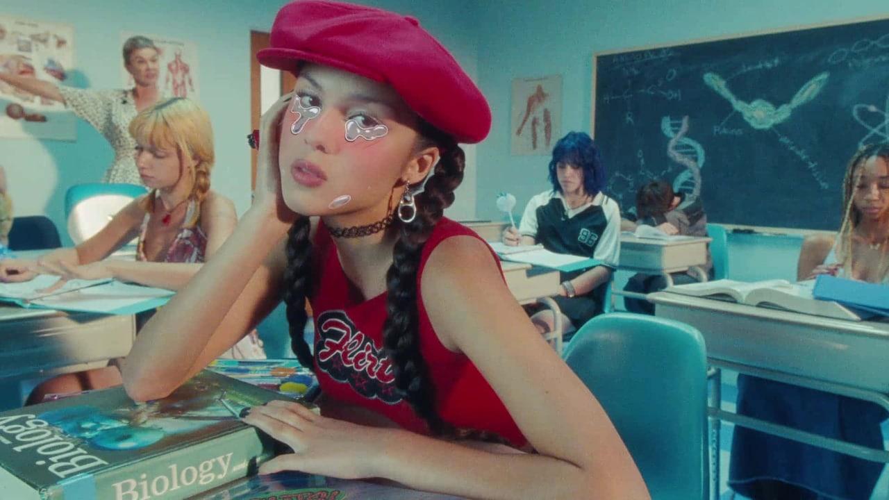 Olivia Rodrigo sits at a school desk in a red beret amid a staged classroom. Other students, books and chalkboard drawings remain at the edges.