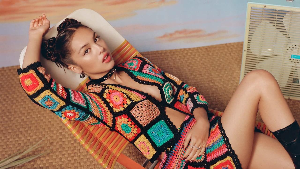 Olivia Rodrigo reclines in a colorful crochet outfit beside a striped chair and box fan. The patterned clothing and room props frame the warm portrait.