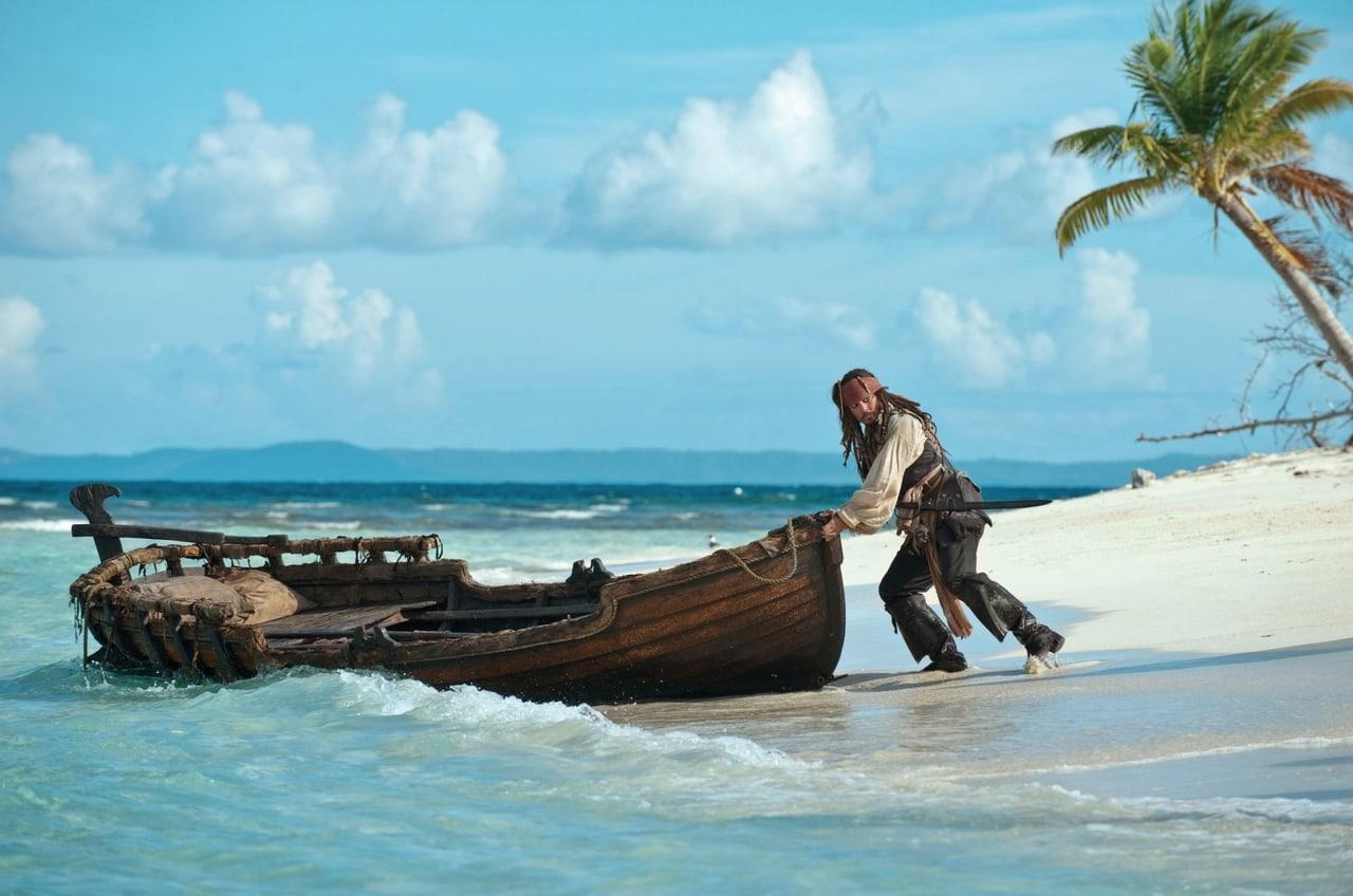Jack Sparrow pulls a weathered boat across a turquoise shoreline beneath a leaning palm. Surf and sunlit sand open the scene out around him. Source: WallpaperCat (uploader wzieme). Original artist not identified.