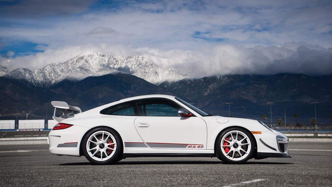 A white 2011 Porsche 911 GT3 RS 4.0 stands in side profile on a circuit with snow-covered mountains beyond.