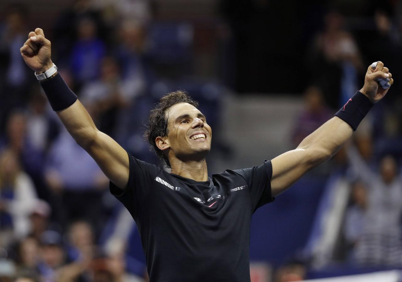 Nadal celebrates in a black shirt with both arms raised. His fists and blurred spectators frame the conversation against the dim arena background.