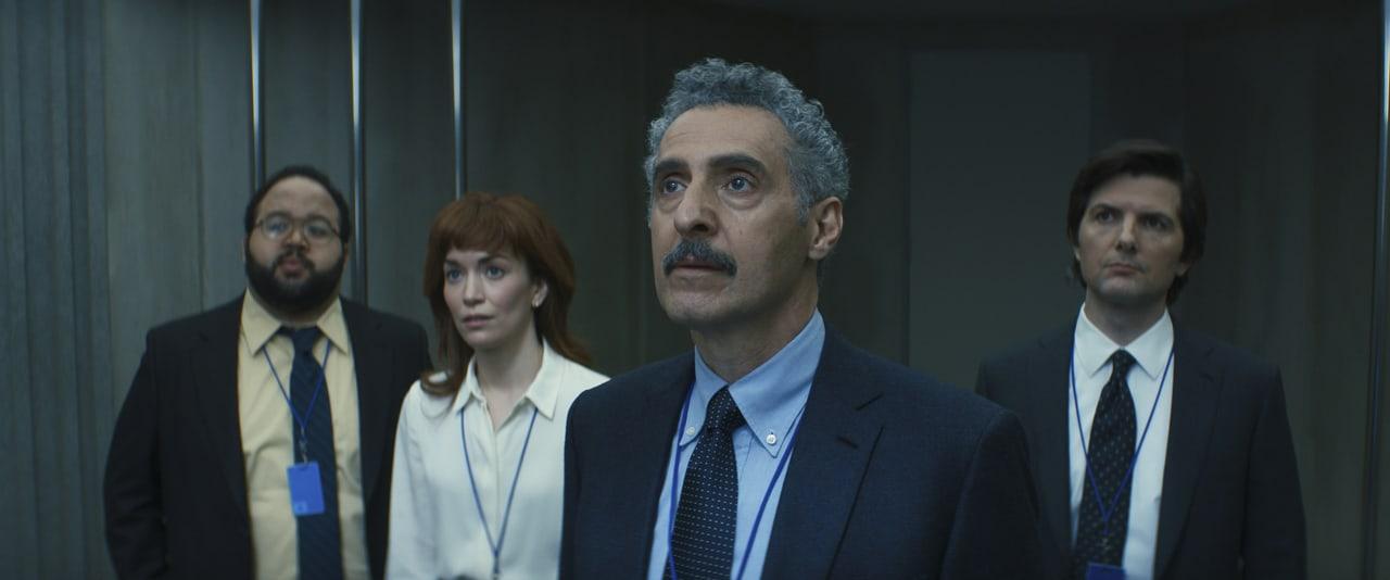 Four colleagues wait inside a steel elevator, with Dylan and Mark holding the outer edges of the frame. Cold metal and muted suits establish the uneasy mood. Source: WallpaperCat (uploader ernser.ariane). Original artist not identified.