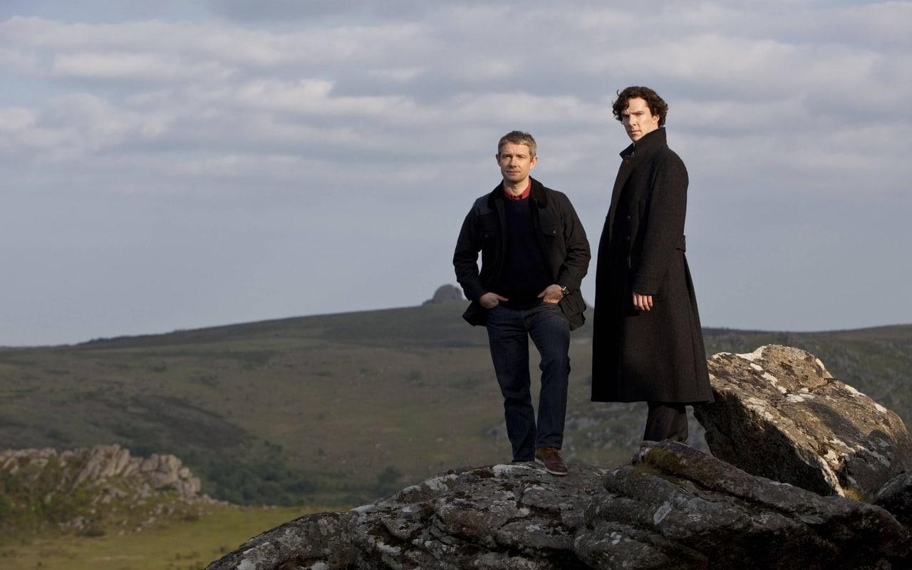 Sherlock and Watson stand on a rocky outcrop under a gray sky. Rolling hills and broken stone continue at both sides of the central pair.