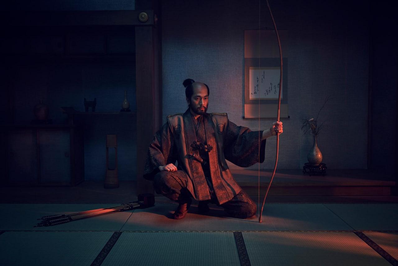 A seated archer holds a bow in a dim room lit with blue shadows and a narrow amber glow. Tatami lines and display alcoves establish the period setting. Source: Alpha Coders, discovered by patrika.