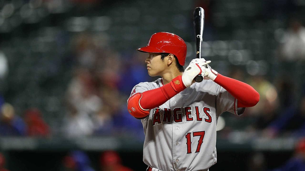Ohtani holds his bat ready in a historical gray Angels uniform. Dark rows of seats and spectators stretch across the background around the central batting stance.