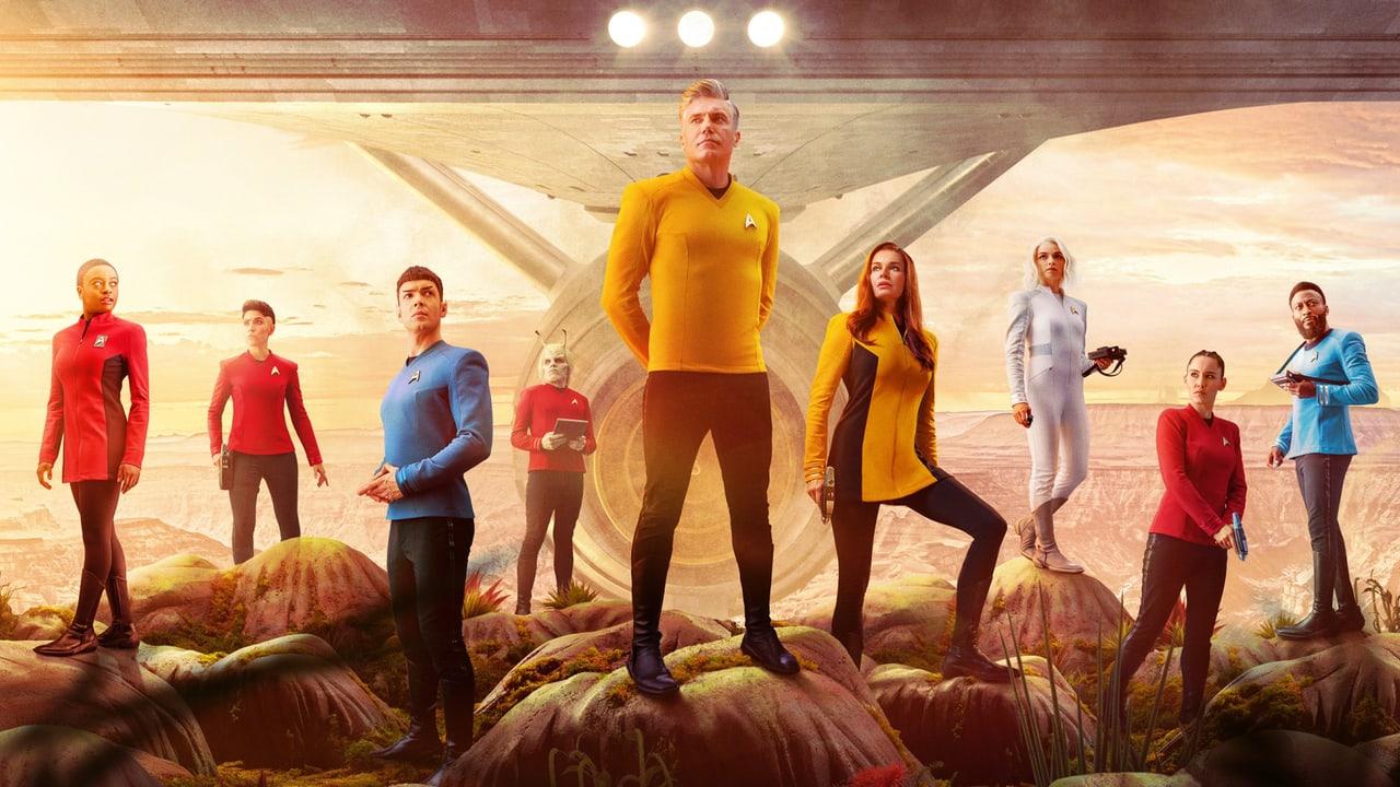 The Strange New Worlds crew stands among alien plants below a starship. Red and blue uniforms at the edges continue the ensemble beyond the central command officers.