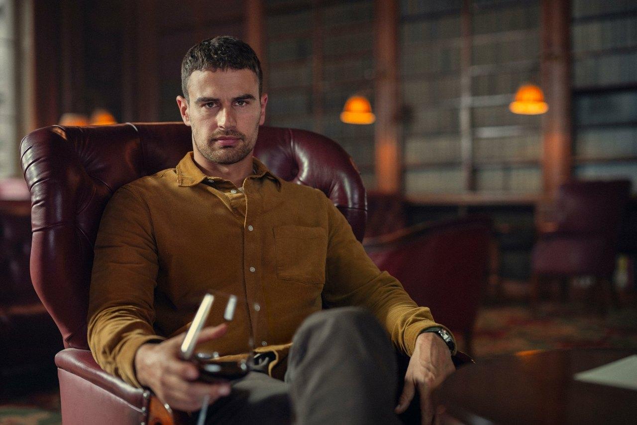 Theo James sits in a burgundy leather chair, wearing an ochre shirt in a dim wood-lined room.