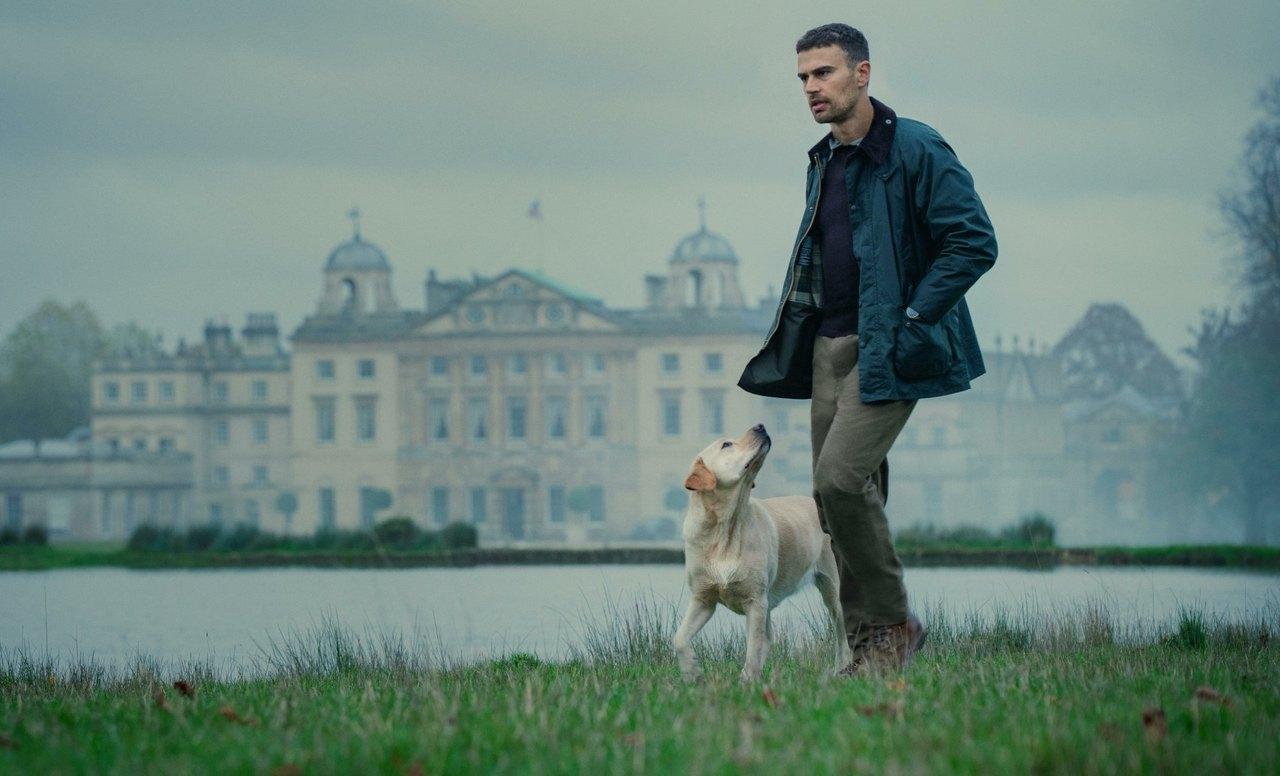 Theo James as Eddie stands beside a dog on the lawn in front of a large country house under a pale sky.