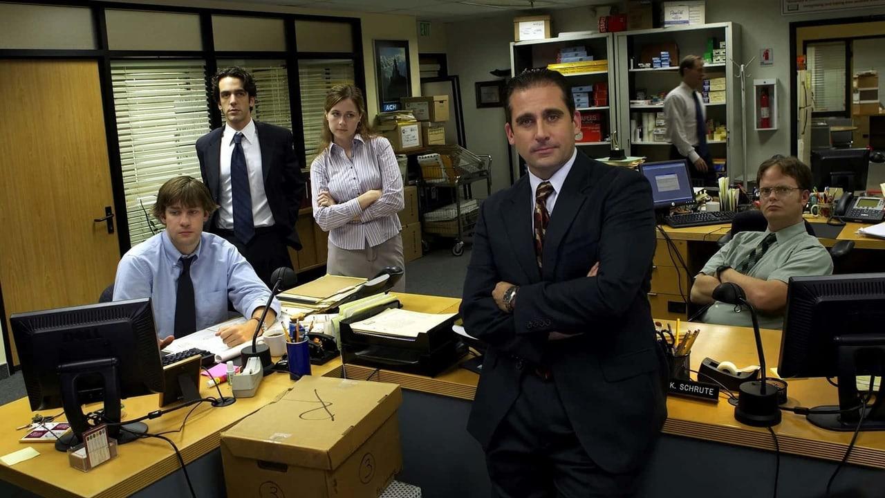 The Dunder Mifflin staff pauses among computers, folders and yellow-topped desks. A monitor and door frame the left while Dwight and office shelves remain on the right.