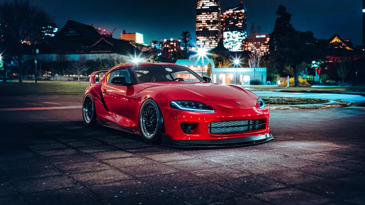A red A90 Supra sits in a city park after dark among traditional rooftops and illuminated towers. Streetlights and reflected color broaden the urban setting. Source: 4kwallpapers.com. Original artist not identified by the source page.