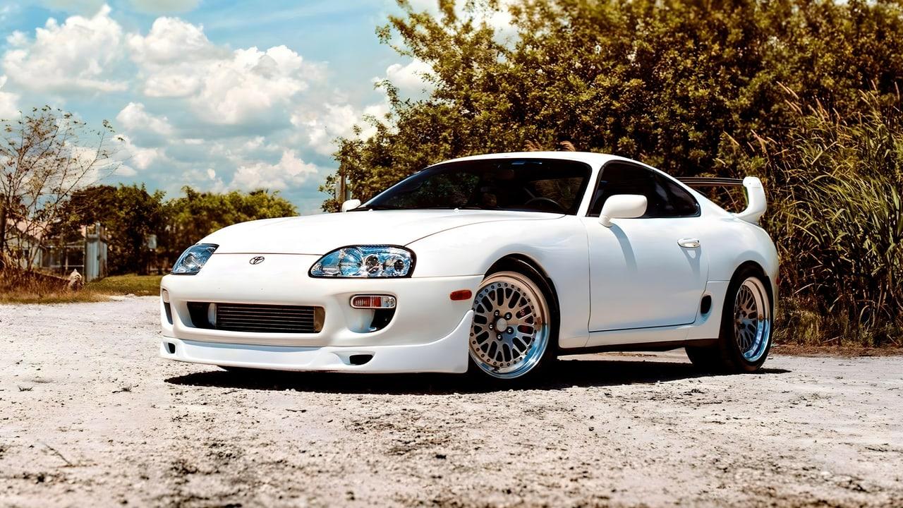 A white Mk4 Supra rests on pale gravel beside trees beneath a cloud-filled sky. Vegetation and the open turnout surround the rounded bodywork. Source: 4kwallpapers.com. Original artist not identified by the source page.