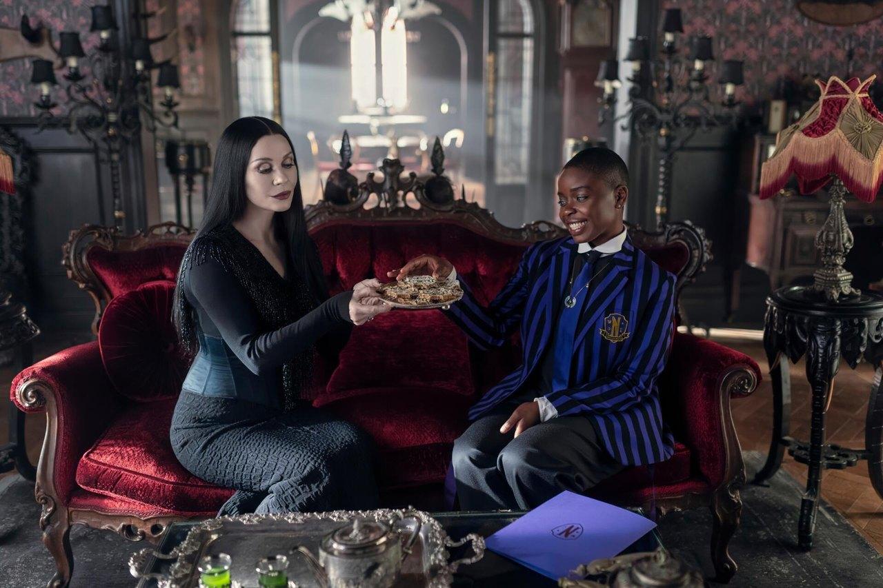 Two women sit on a red velvet sofa amid carved dark furniture, candles and a fringed lamp.