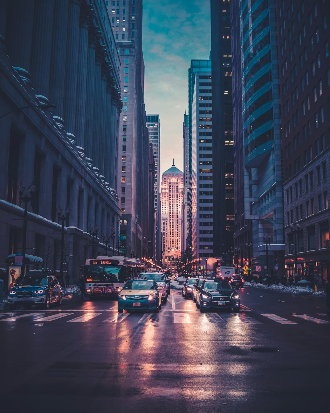 A moody city street scene shot from the center of a wide avenue lined with tall buildings. The perspective draws the eye toward a glowing focal point in the distance, where streetlights and vehicle headlights create warm amber accents against predominantly cool teal and navy tones. The roadway appears slightly reflective, adding subtle shimmer and depth. The overall lighting suggests dusk or early night, with a cinematic contrast between dark architecture and illuminated windows. Composition is symmetrical and immersive, evoking a quiet, urban atmosphere with a sense of scale and anticipation.