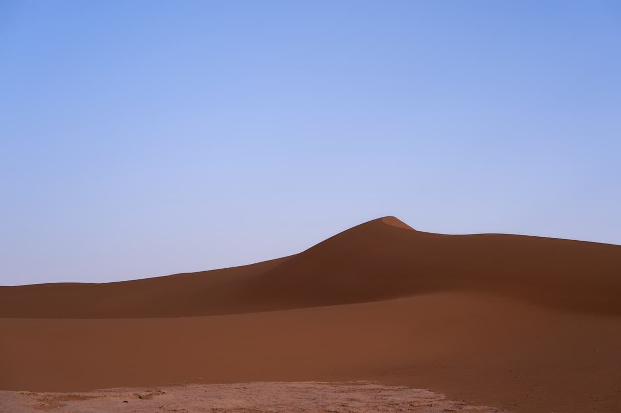 Simple landscape photograph of a single dune ridge stretching horizontally across the frame. The sand is a rich brown tone, smooth and unbroken, beneath a clear gradient sky that shifts from lighter blue near the horizon to deeper blue overhead. The foreground is flat and lightly textured, while the dune's crest forms a clean, minimal silhouette. Lighting is soft, with subtle shading rather than harsh contrast. The overall composition is understated and balanced, emphasizing calm and emptiness.