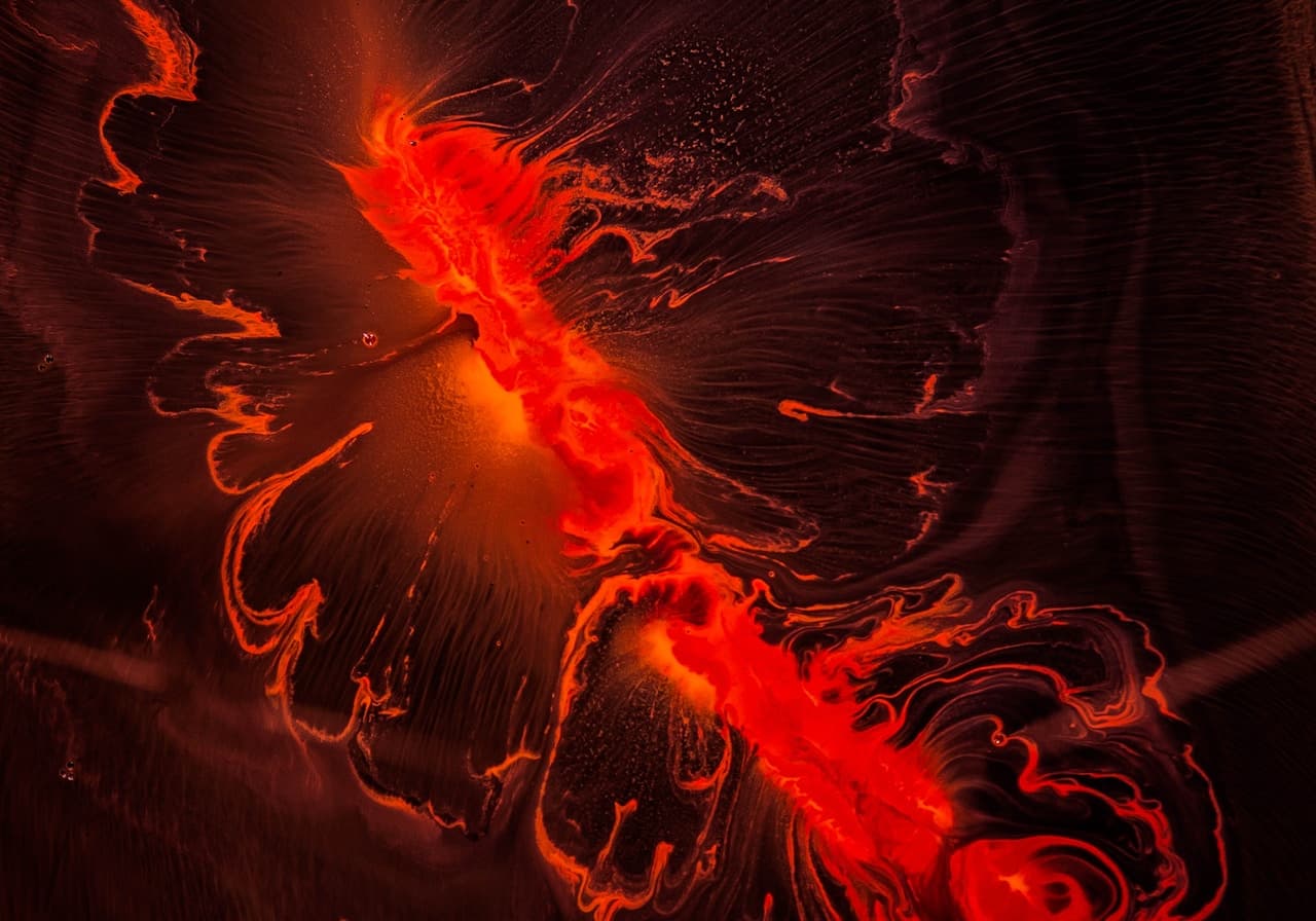 A moody close-up image dominated by a vivid red-orange form against a nearly black background. The subject reads like a macro of flowing pigment or flower-like petals, with glossy highlights and fine ripples that create a sense of movement. Dark negative space surrounds the main shape, keeping the composition minimal and dramatic. Lighting is directional and soft, catching textured edges and creating deep shadows. The overall atmosphere is intense and intimate, with a fiery, abstract mood.