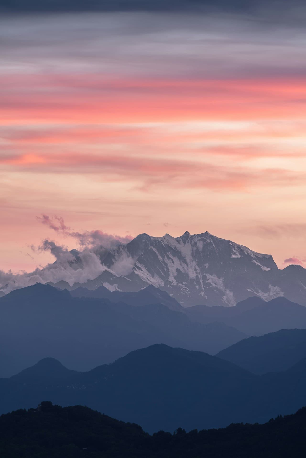 A vertical landscape photograph of a snow-covered mountain rising behind darker, layered ridgelines. Above, soft pink and orange clouds glow against a cool blue sky, suggesting sunrise or sunset. The composition stacks the ridges in receding layers, creating strong depth and a sense of scale. The light is gentle and atmospheric, with pastel tones in the sky and cooler shadows on the slopes. The overall mood is calm and majestic, blending crisp alpine detail with a serene, painterly color gradient in the sky.