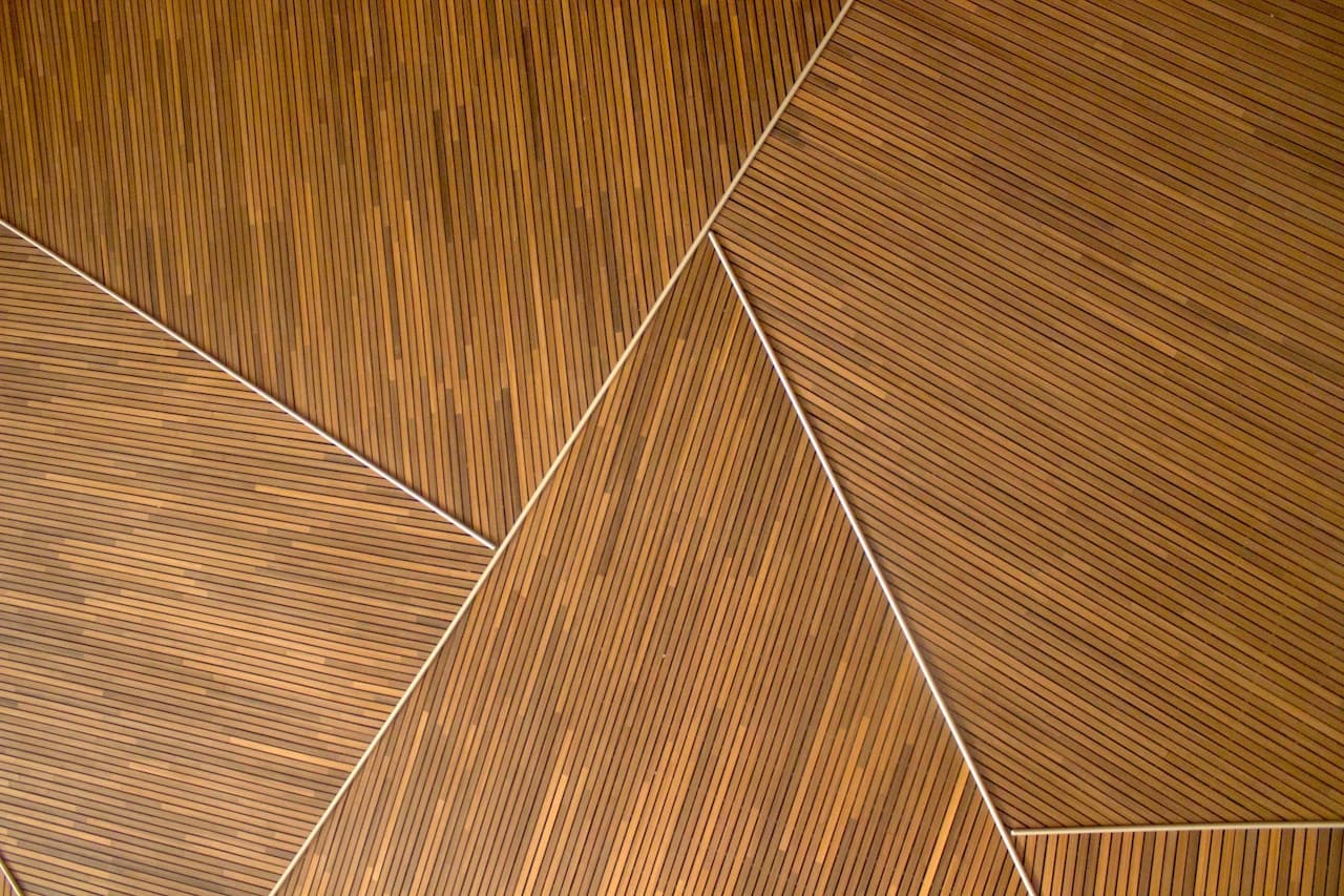 Geometric close-up of wooden panels made from narrow, evenly spaced slats. Multiple panels meet at sharp angles, creating triangular and polygonal shapes outlined by thin light seams. The warm wood grain and repeating lines produce a strong sense of rhythm, while subtle variations in tone suggest different planks or lighting across surfaces. The framing is tight and abstracted, turning a ceiling or wall detail into a pattern study. Lighting is warm and even, with minimal shadows. Mood is cozy, modern, and architectural.